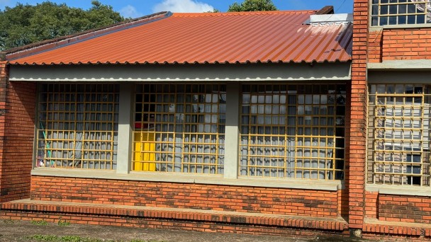 Imagem de prédio da escola, feito em tijolo à vista. A maior parte é ocupada por uma construção de um andar, mas na ponta direita vê-se a continuação desta, com dois andares.