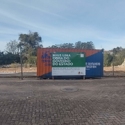Em frente a um contâiner metálico, uma placa anuncia a obra do estado, com dois homens de capacete de obra laranja na esquerda.