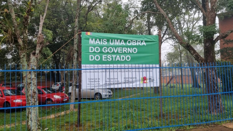 Placa anuncia obra do governo do Estado atrás de uma cerca azul e em meio a árvores. Ao fundo aparecem carros no estacionamento.
