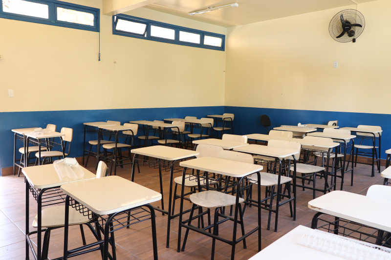 Sala de aula com paredes pintadas em azul embaixo e amarelo em cima, com classes e cadeiras e um ventilador de parede ao fundo.