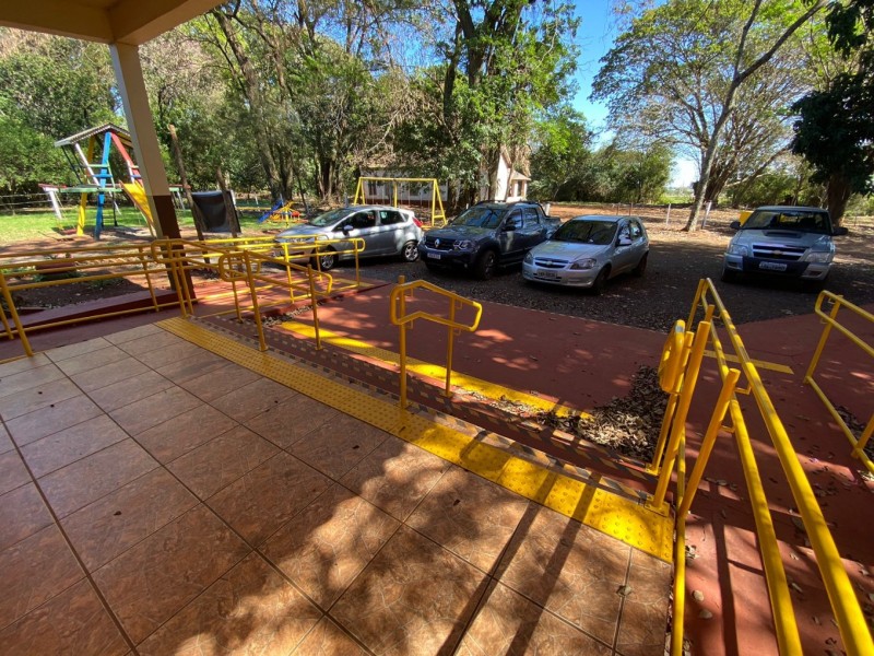 Entrada da escola com rampa de acesso e corrimãos, com quatro carros estacionados à frente e parque de diversões ao lado.