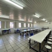 Refeitório de escola, com paredes, piso, teto, cortinas, mesas e cadeiras brancas.