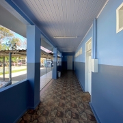 Corredor externo da escola, pintado em dois tons de azul, com quadra de vôlei de cimento com crianças jogando ao fundo.