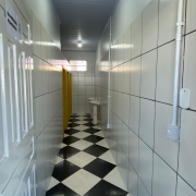 Entrada de banheiro com piso quadriculado em preto e branco, paredes, teto e porta brancos e duas pias e duas portas ao fundo.