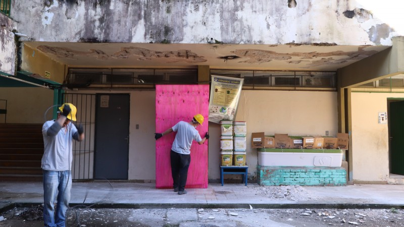 Parte interna de escola com dois homens de chapéu de obra amarelos trabalhando.