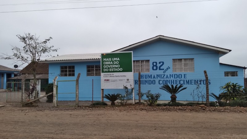 Prédio da escola em terreno de chão batido. Prédios pintados em azul claro, com uma placa indicando a obra na frente.