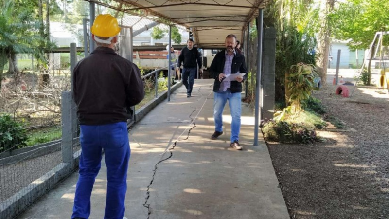 Em um corredor externo coberto, três homens circulam, um deles com capacete de obra amarelo, tendo um parque infantil à direita.