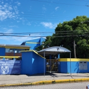 Faixada frontal da escola Luiz Moschetti, pintada em azul e amarelo, com carro e poste à direita.