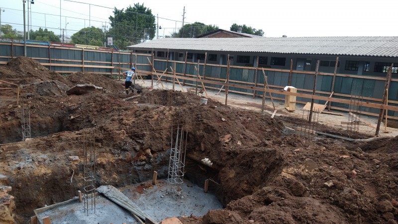 Obra de construção de prédio com buracos para as fundações, um homem com um carrinho de mão ao fundo.