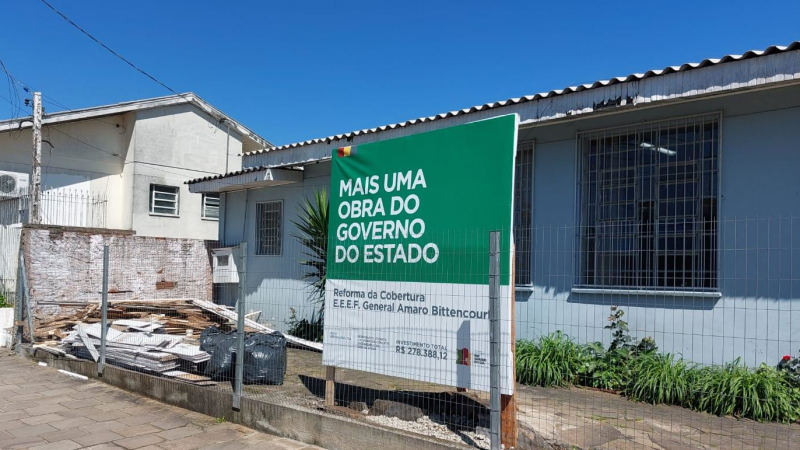 Parte frontal da escola, com prédio ao fundo e ao lado uma casa, e à frente, atrás da cerca, placa indicando a obra e restos de madeira.
