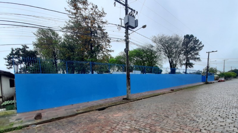 Em primeiro plano, uma rua de calçamento de pedra com um carro vermelho ao fundo. Atrás, muro em azul e postes de luz.