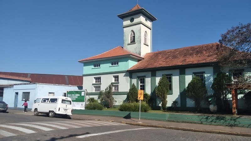 Em primeiro plano, uma rua de calçamento de pedra com uma faixa de pedestres. Ao fundo, uma Kombi branca na frente do prédio da escola, pintado em tons de verde e com uma torre.