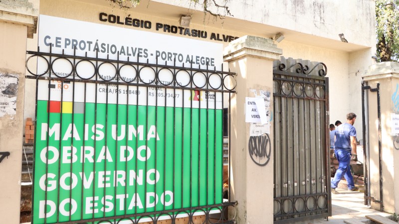 Em primeiro plano, muro e grade da entrada da escola com placa anunciando a obra. Atrás, um homem vestido de azul passa com um carrinho de mão. Ao fundo, o letreiro com o nome da escola.