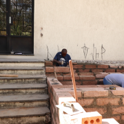 Escada à esquerda e, ao centro e à direita, rampa de acessibilidade sendo construída com pedras. Dois homens trabalham na rampa.