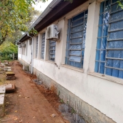 Parte externa da escola. À esquerda, duas árvores. Ao centro e à direita, o prédio escolar. No chão, placas do piso.