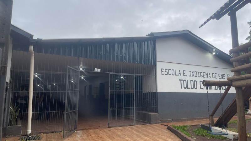 Parte frontal da escola, pintada em azul e branco, com céu nublado e parte de um parquinho à direita.