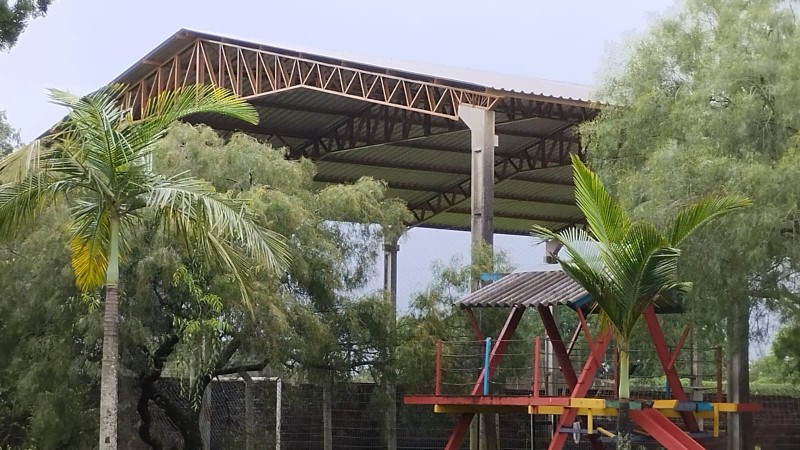 Atrás de árvores e um parquinho, a estrutura da cobertura da quadra de esportes.