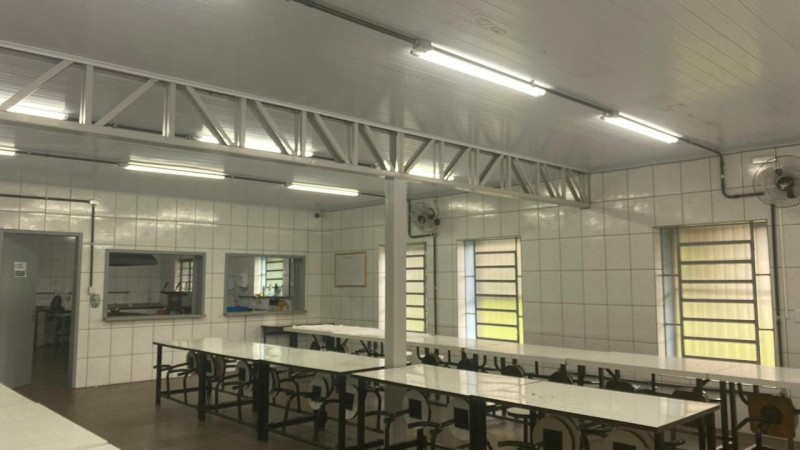 Parte interna de refeitório, com paredes brancas e duas mesas longas com cadeiras.