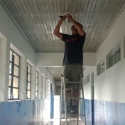 Num corredor com as paredes pintadas em azul, abaixo, e branco, acima, com janelas do lado esquerdo e salas de aula do lado direito, um homem em cima de uma escada trabalha em uma instalação no teto.