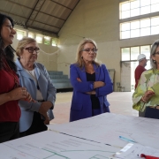 Dentro do ginásio da escola, quatro mulheres estão em volta de uma mesa onde se vê a planta do projeto. Ao fundo, dois operários conversam.