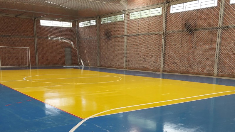 Interior de ginásio, com quadra pintada em amarelo e azul e paredes em tijolo à vista.