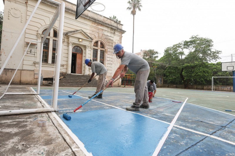 Três homens pintam o piso de uma quadra poliesportiva.