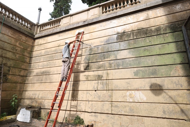Homem em uma escada utiliza um lava-jato para limpar um muro.