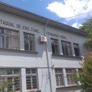 Vista frontal do prédio de dois andares da escola, pintada em cor cinza, com árvores à direita.