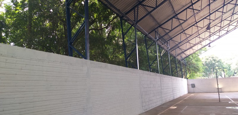 Vista interna da quadra coberta, com destaque para o muro branco e a cobertura em metal azul e telha metálica. Ao fundo, veem-se árvores.