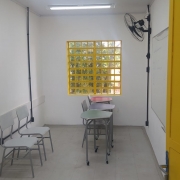 Vista interna de uma sala de aula. As paredes são brancas e a janela e a porta amarelas. Há um quadro branco e um ventilador na parede e duas cadeiras e três mesas na sala.