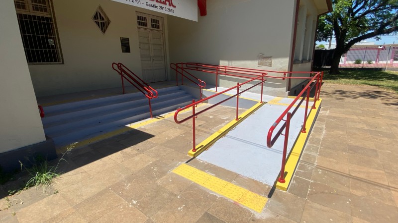 Foto da entrada do prédio da escola com rampas e escada com corrimãos.