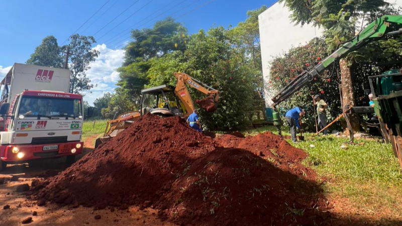 Atrás do prédio da escola, uma patrola revira a terra, tendo um caminhão à esquerda e quatro homens à volta.
