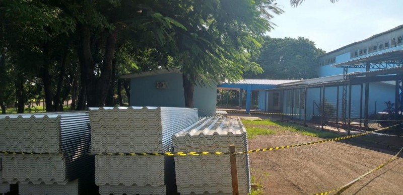 Em primeiro plano veem-se telhas e uma fita de isolamento. Ao fundo, árvores e o prédio escolar em azul claro.
