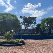 Vê se a rua de pedras, com um canteiro florido no centro, contando com um letreiro com o nome da escola. Ao fundo, o prédio escolar.