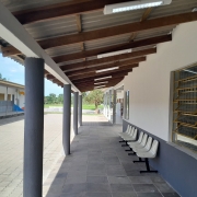 Vê-se o pátio da escola, com um corredor coberto à frente e um dos prédios à direita e outro à esquerda.