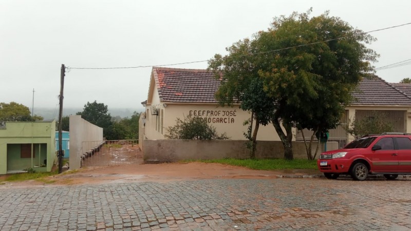 Em primeiro plano, o calçamento de pedras. Ao fundo, à esquerda, uma casa verde e, atrás, outra azul. No centro e na direita, o prédio da escola, com uma árvore e um carro vermelho à frente.