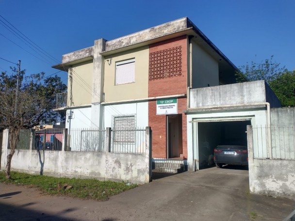 Imagem de uma casa de dois pisos com parte da parede em tijolo à vista e parte pintada em azul claro no andar de baixo e amarelo escuro no andar de cima. À direita, uma garagem com um carro azul escuro estacionado. À esquerda, fora do pátio, uma árvore pequena.