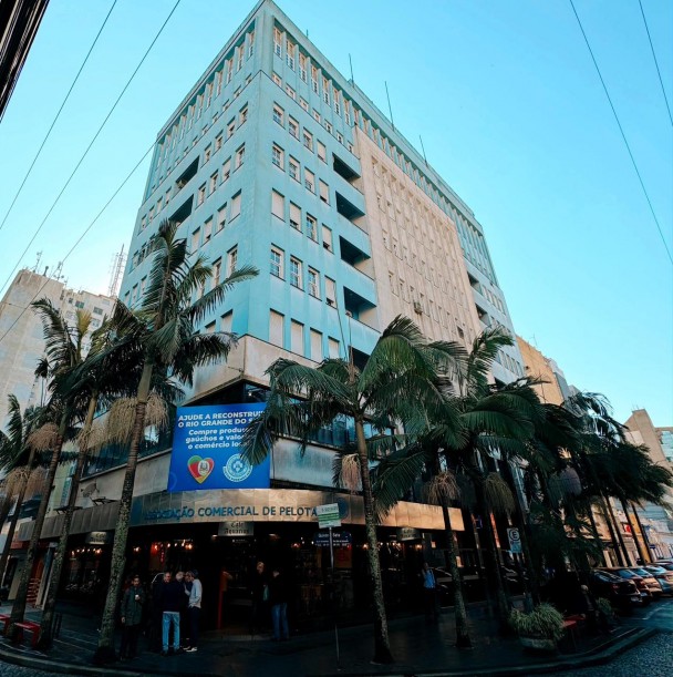 Imagem de um prédio nas cores azul e amarelo. Há pessoas em frente e um letreiro dizendo Associação Comercial de Pelotas. À frente do prédio, há palmeiras. Uma placa azul aparece em cima do letreiro. O céu está azul.