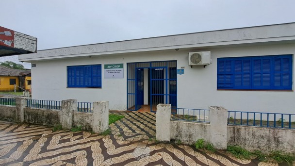 Imagem de uma casa de um piso pintada em bege com as janelas azuis. O céu está nublado. À esquerda, veem-se parte de uma casa amarela e a cobertura de uma parada de ônibus. À frente da casa, gramado e uma cerca baixa. Na parte inferior da foto, a calçada.