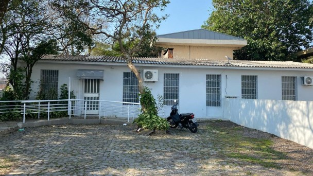 Imagem de uma edificação de um piso branca, com uma árvore e uma motocicleta na frente e um muro branco do lado direito.