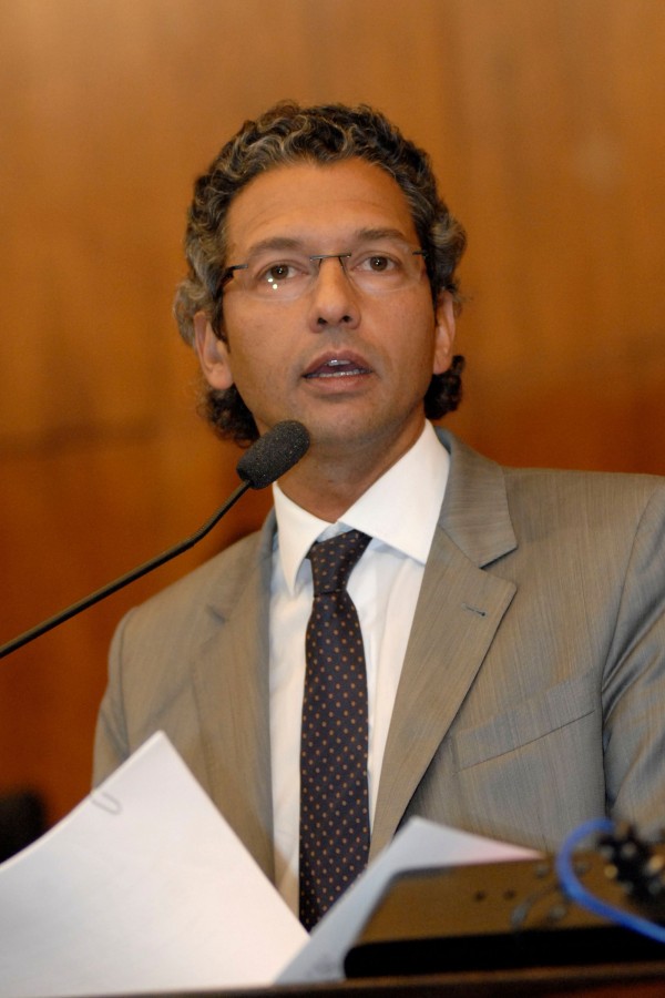 Foto de um homem falando à tribuna com um papel à frente e um microfone.