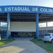 Foto da fachada da escola. Há uma área coberta onde se lê "Escola estadual de Colinas". À frente, uma calçada entremeia gramados, onde estão estacionados quatro carros, dois de cada lado.