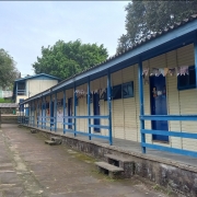 Foto do pátio interno de uma escola. O piso é de pedra, à direita um prédio de um piso, de madeira, pintado em amarelo e azul. Ao fundo, à esquerda, um prédio de tijolos com uma pintura com o nome da escola e, à direita, a continuação, no alto, do prédio de madeira.