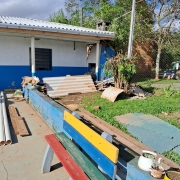 Foto do pátio da escola, com parte do prédio ao fundo, pintado em branco e azul, com uma churrasqueira nas mesmas cores. No pátio, veem-se, pelo chão, canos e telhas.