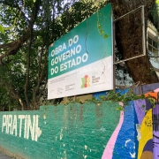 Foto do muro frontal da escola, pintado em verde. Lê-se, nele, a palavra "piratini". Acima dele, árvores e a placa indicando uma obra do governo do estado.