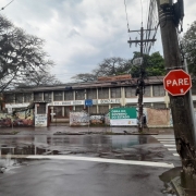 Foto externa da fachada da escola. Em primeiro plano, um poste com uma placa de pare. Em sequência, a rua, molhada. Ao fundo, o prédio de dois pisos da escola, com uma placa indicando a obra do estado no muro frontal. O céu está nublado.