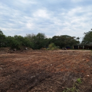 Vê-se um campo aberto com terra mexida. Ao fundo, árvores e um prédio de um piso. O céu está azul com nuvens.