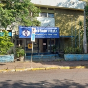 Foto externa do prédio escolar de dois pisos. Vê-se a rua asfaltada em primeiro plano e, na calçada de pedras, uma placa de estacionamento com vaga para deficiente. Na entrada da escola há um letreiro azul com o nome da instituição.