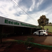 Vê-se o prédio de um piso da escola, pintado em verde. À esquerda, o letreiro traz o nome da escola. O céu está azul com nuvens. Há árvores em volta do prédio. A rua é de brita, há um carro branco estacionado e duas pessoas aparecem, uma caminhando e outra sentada.