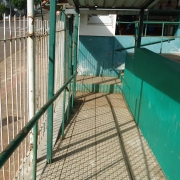 Foto tirada dentro do terreno da escola. Há um piso em plano mais alto à direita. Ao centro, a rampa de entrada da escola. As muretas são verdes. À esquerda, a cerca branca e a parte externa, com uma rua onde se vê um carro.